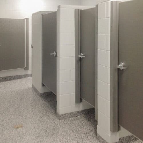 Photograph of a public restroom showcasing four SL-175 bathroom stall doors, white tiled walls, and gray speckled flooring. Each stall door is fitted with a sturdy metal handle.