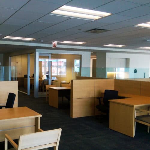 An office space with wooden desks, chairs, and partitions, featuring overhead fluorescent lights and a glass-enclosed meeting room in the background. The area appears clean and organized.