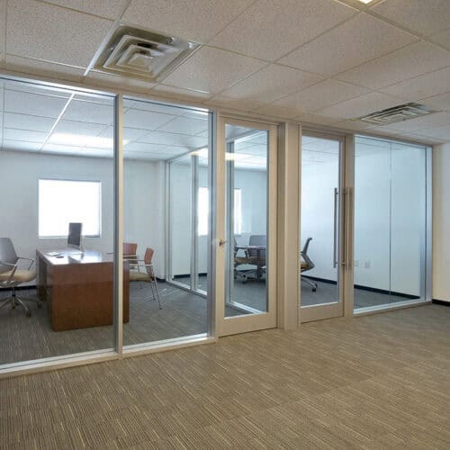 An empty office with glass-walled meeting rooms, modern furniture, and gray carpeting.