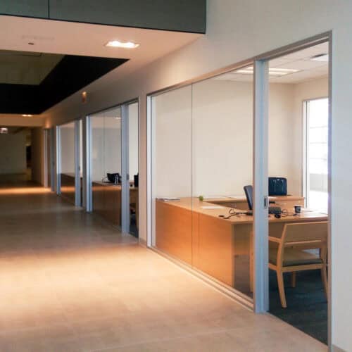 A hallway with glass-enclosed office cubicles on the right, each containing desks, chairs, and office equipment. The area is well-lit with artificial and natural light.