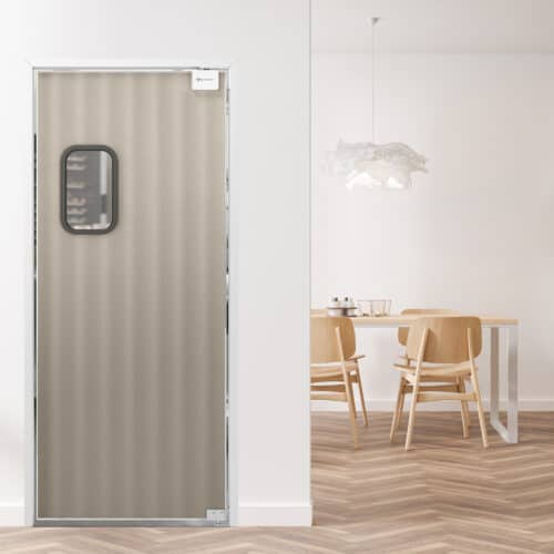 A beige textured traffic door with a small rectangular window is situated in a white room. In the background, there is a dining table with four wooden chairs and a decorative hanging light fixture.