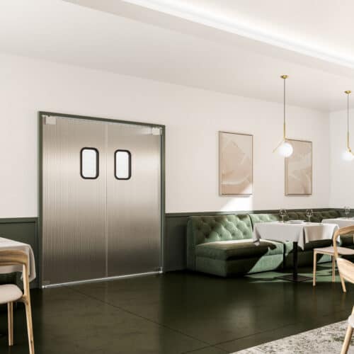 A modern dining area featuring green upholstered seating, two white tables set with glassware, pendant lights, and double traffic doors with small windows.