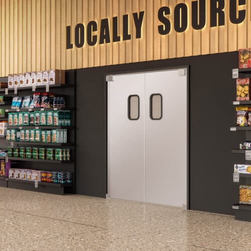 A grocery store aisle featuring shelves stocked with various products. A set of double traffic doors with small windows is seen on the rear wall beneath a “Locally Sourced” sign.