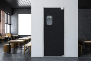 A black traffic door with a small window, set in a white wall, leads to an area with several wooden tables and chairs arranged in a modern, dimly lit interior space.