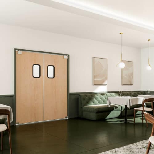 A modern, minimalist dining area with green cushioned seating, wooden tables, and pendant lights. Double wooden traffic doors with small windows are in the background. Light streams through a window on the right.