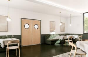 A modern, minimalistic dining area with wooden tables, green cushioned seating, pendant lights, and double traffic doors with porthole windows. Light streams through a large window to the right.