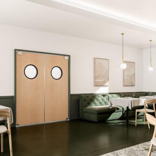 A modern, minimalistic dining area with wooden tables, green cushioned seating, pendant lights, and double traffic doors with porthole windows. Light streams through a large window to the right.