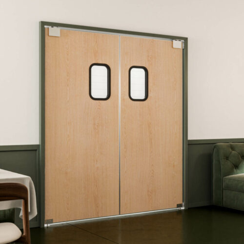 A modern, minimalist dining area with green cushioned seating, wooden tables, and pendant lights. Double wooden traffic doors with small windows are in the background. Light streams through a window on the right.