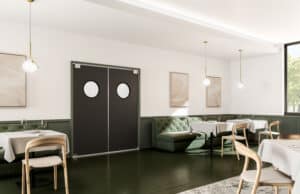 A modern restaurant interior featuring green tufted seating, wooden chairs, white tablecloths, and a pair of double doors with porthole windows. The room has minimalist decor and pendant lighting.