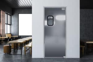 A modern, empty dining area with wooden tables and chairs to the left, featuring a closed metal door with a small window in the center background wall.