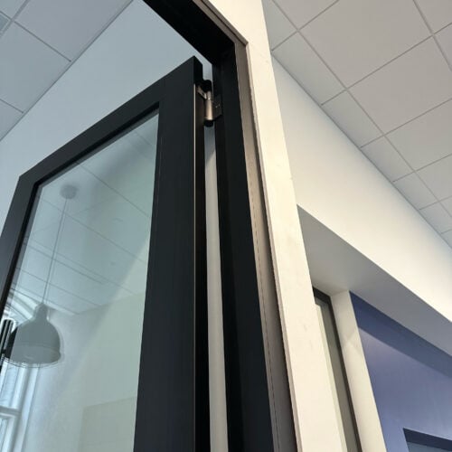 A black-framed glass door is partially open, revealing a hinge and a white wall. The ceiling has white tiles, and a hanging light fixture is visible in the reflection on the glass partition walls with enhanced privacy.