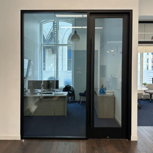 Glass-walled office with a closed door, featuring a desk, dual monitors, office chair, and a lamp. Background shows part of another room and an outdoor city scene visible through the glass partition walls with enhanced privacy.