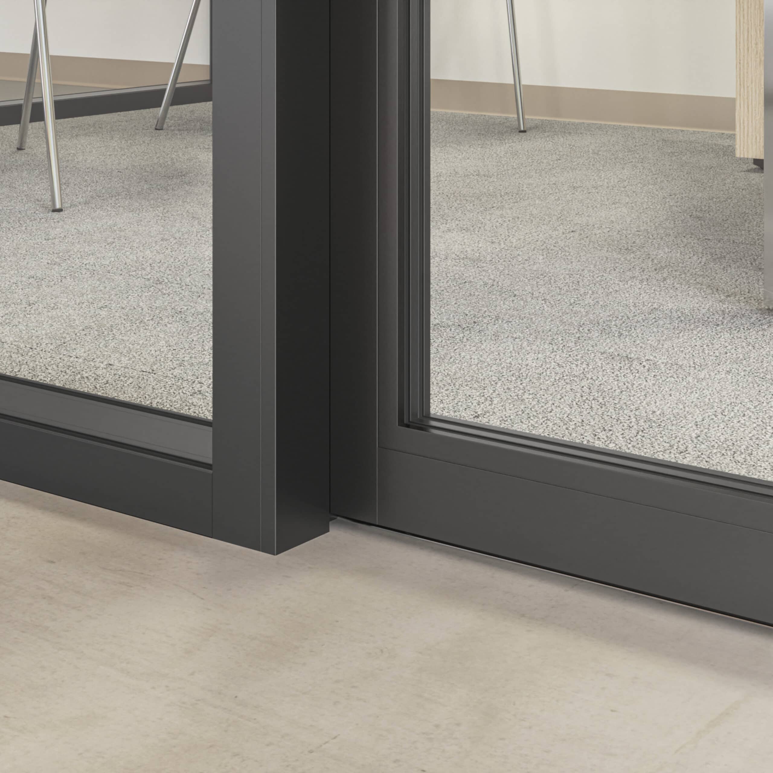 Close-up of a modern glass door with a black frame, revealing part of a carpeted office space inside, including chair legs and a wooden desk. The floor outside the door appears to be tiled, highlighting the sleek transition provided by glass partition walls with enhanced privacy.