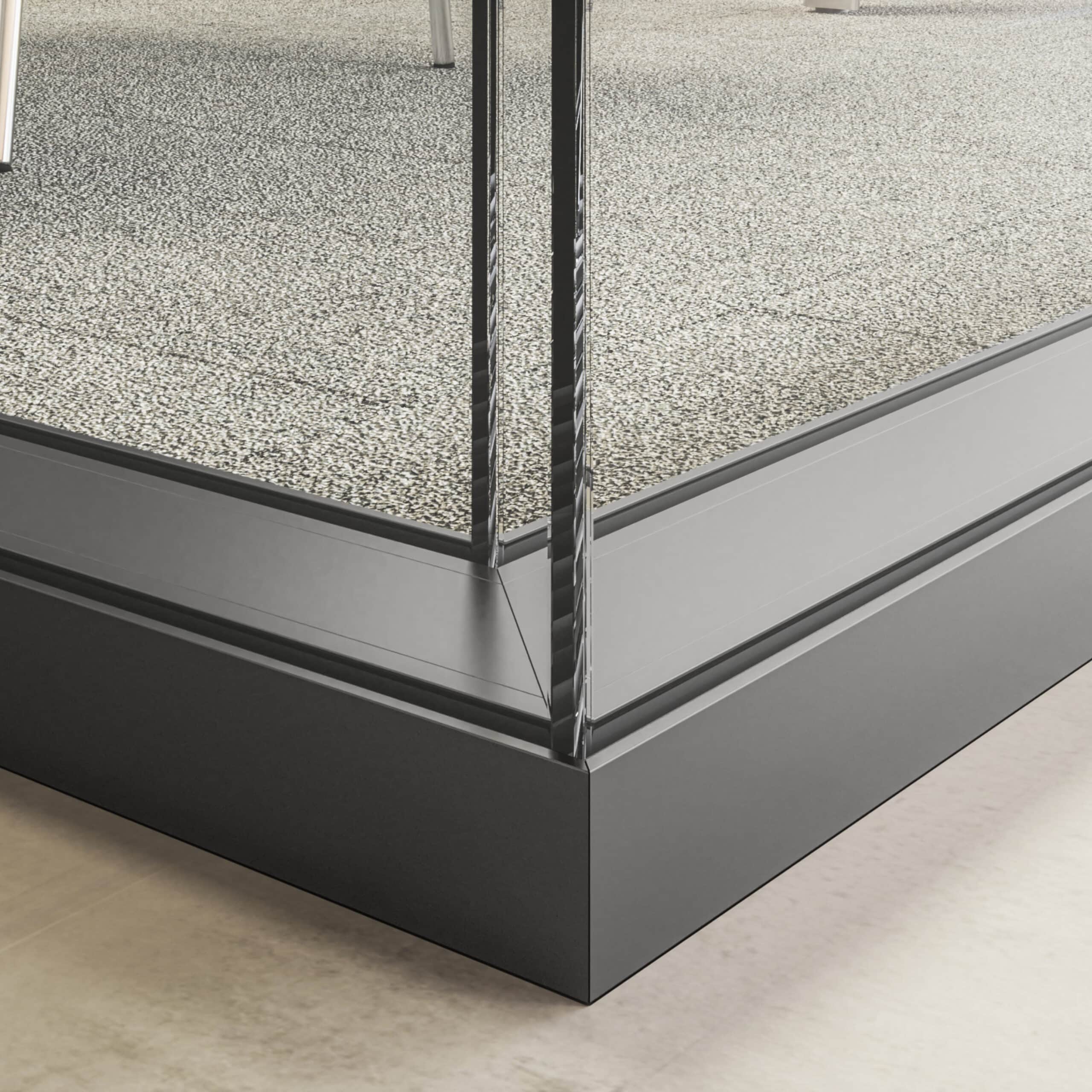 Close-up of the corner of a glass partition wall with enhanced privacy, framed in metal and mounted on a gravel-covered surface, with a smooth, light-colored floor on the bottom left. The corner is reinforced for stability.