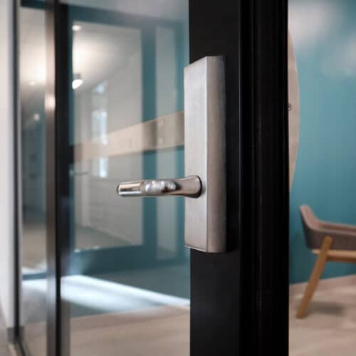 Close-up of a metallic door handle on a glass door. In the background, there is a blue wall and a wooden chair with a gray cushion, part of an office space featuring glass partition walls with enhanced privacy.