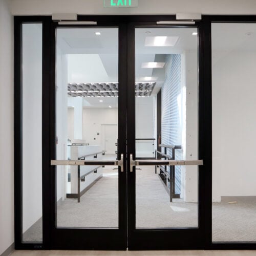 A set of double glass doors with black frames in a modern building interior, leading to a hallway with white walls and a visible EXIT sign above, complemented by adjoining glass partition walls with enhanced privacy.