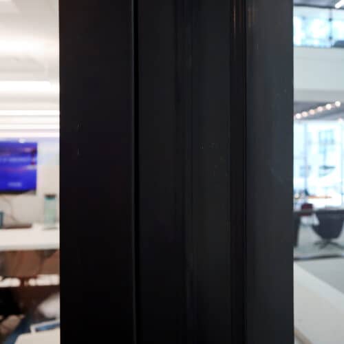 Close-up of a dark vertical divider with an out-of-focus office space background featuring desks, chairs, and a screen on the left side, enhanced by glass partition walls offering improved privacy.