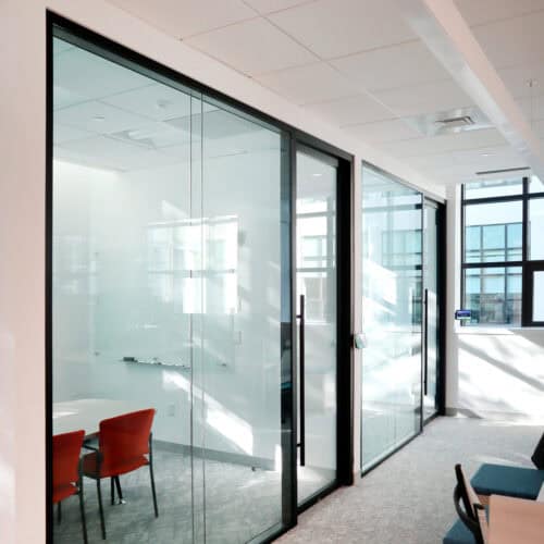 A modern office space features glass partition walls with enhanced privacy, glass-enclosed meeting rooms, red chairs, white walls, and carpeted floors. Natural light enters through large windows.