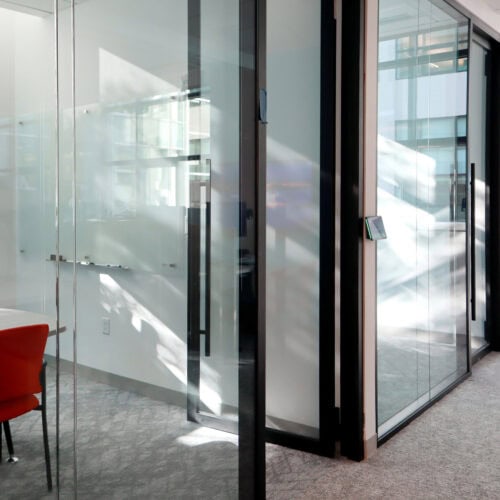 A modern office space with glass partition walls with enhanced privacy features, a red chair, and a small digital device on one of the walls. Sunlight streams through the windows, casting reflections and shadows on the floor and walls.