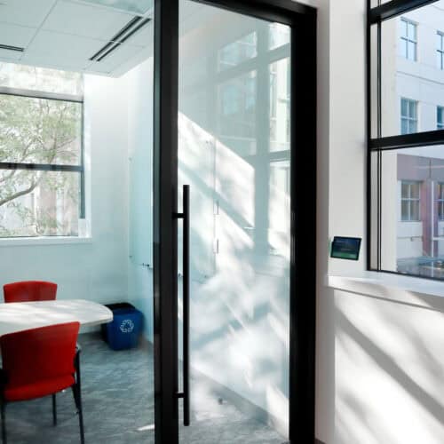 Glass-walled conference room with a round table, red chairs, and a recycling bin. Sunlight streams through the windows, creating shadows on the walls and floor. The glass partition walls with enhanced privacy ensure confidential discussions amidst an open ambiance.