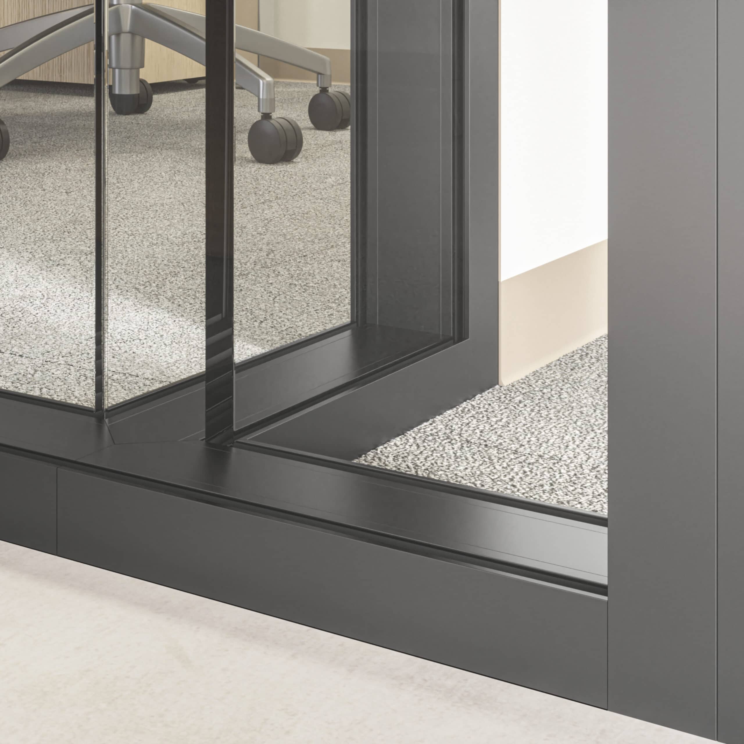 Close-up of the lower section of glass partition walls with enhanced privacy featuring a black aluminum frame. Rollers of an office chair are visible through it, and the floor is covered with grey carpet tiles.