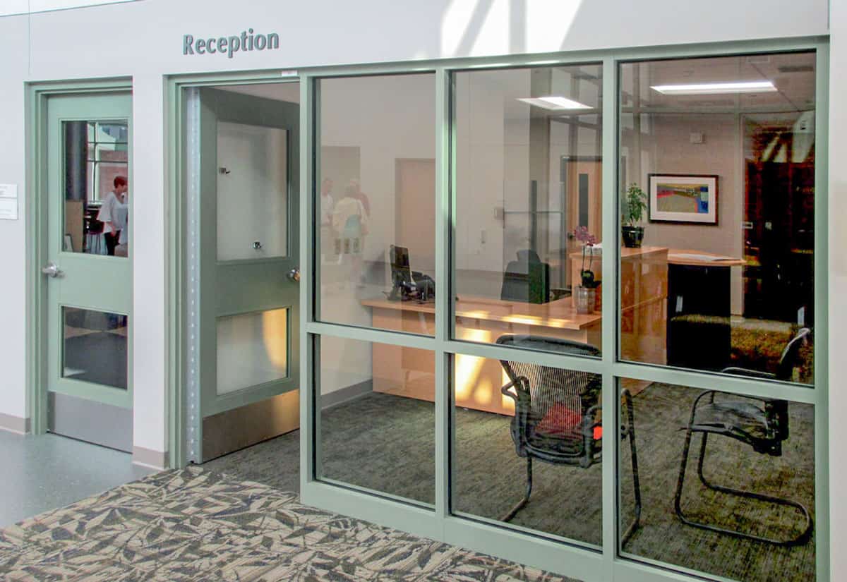 A reception area with a glass window, reception desk, chairs, and a printer provides a welcoming atmosphere for visitors seeking healthcare solutions. The open door invites you in, while the carpet's leaf pattern adds a touch of nature.