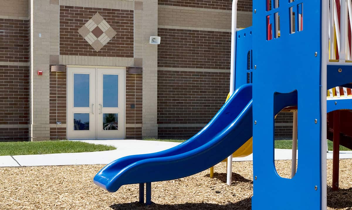 Image of a playground slide beside a building with double doors and a brick wall. The slide is blue and there is wood mulch on the ground surrounding it, ensuring both fun and school security for all the kids.