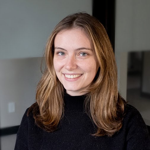 A member of our team, sporting long brown hair and a black sweater, smiles warmly in an indoor setting.