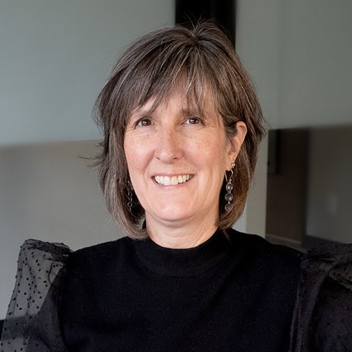 A woman with short gray hair, a key member of our team, wears a black blouse and smiles warmly at the camera against a neutral background.