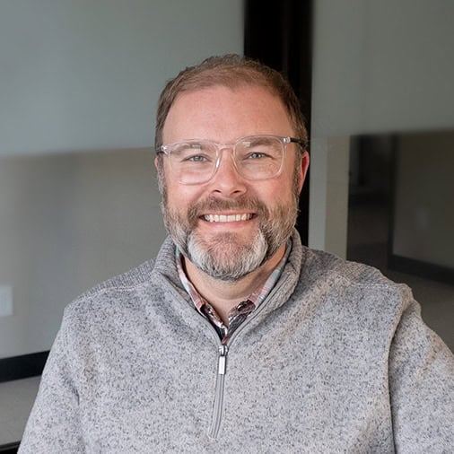 A person with glasses and a beard smiles while seated indoors, wearing a gray zip-up sweater. Our team spirit shines through in every smile.