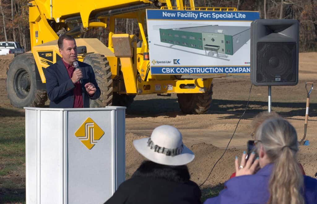 A man speaks at a podium during a groundbreaking event in Decatur. A construction vehicle and a sign for Special-Lite's new manufacturing facility are in the background. People with cameras are taking photos.