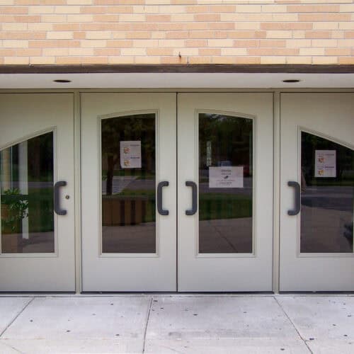 Four smooth aluminum double doors with large glass panes and black handles are set in a brick wall. Signs are posted on the doors.