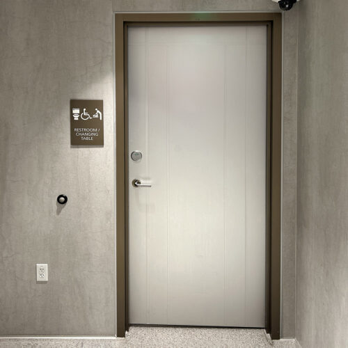 A closed SL-16S restroom door features a sign indicating accessible facilities and a changing table. The sleek surface complements the gray wall, where a light switch is visible.