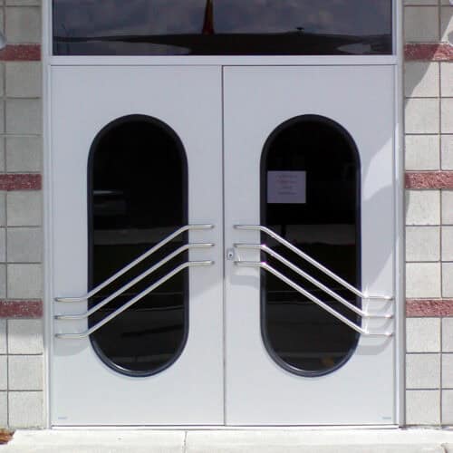 A set of double doors with oval windows, featuring unique wavy metal handles, and a smooth aluminum frame set in a brick wall with horizontal red accents. A small sign is visible on one door.