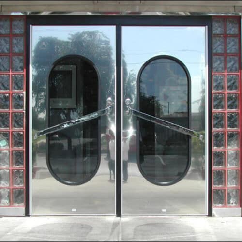 Reflective double glass doors with oval windows and sleek metal handles. Red brick and glass block walls flank the entrance, enhancing the visible outdoor reflections. The stainless steel flush doors add a modern touch to this classic design.