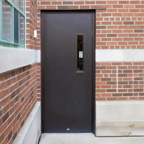 An embossed aluminum flush door with a vertical window is set in a brick wall, next to a smaller glass window.