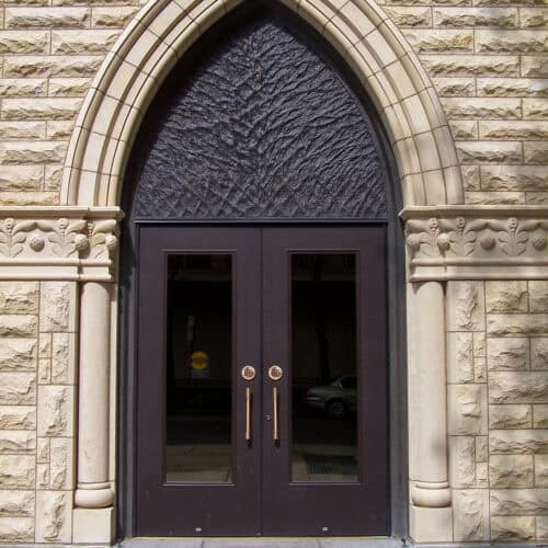 Arched double doors, one embossed aluminum and the other flush, set in a textured stone wall, framed with decorative molding under a pointed arch.