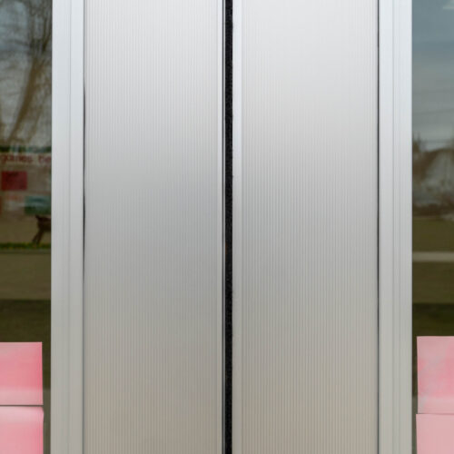 Close-up of a pair of closed, light gray, fluted aluminum sliding doors with panel textures and a narrow black trim in the center. The background is blurry, hinting at outdoor scenery.