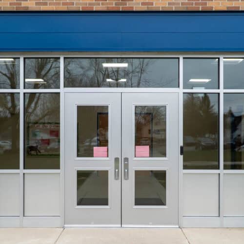 A pair of glass double doors, framed in fluted aluminum, are nestled in a windowed wall. Pink papers are taped to their surface, while fluorescent lights and trees dance as reflections.