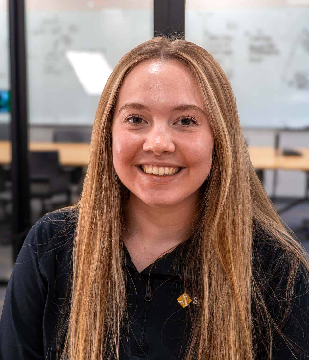 A woman with long blonde hair smiles at the camera, standing indoors in front of glass walls with whiteboards and office furniture visible in the background—capturing the welcoming spirit of our team.