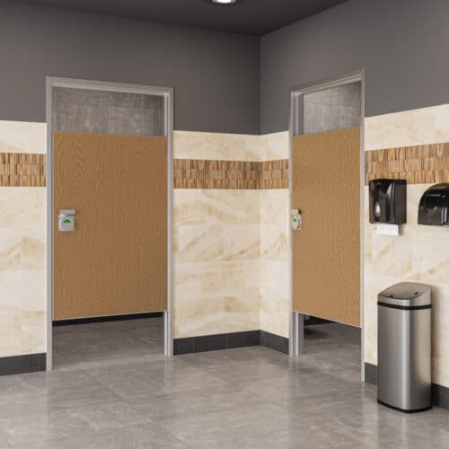 A public restroom with two stalls featuring partial wooden doors set in FlexFit™ Field-Adjustable Interior Aluminum Door Frames, beige tiled walls, a trash can, and paper towel dispensers on the right wall.