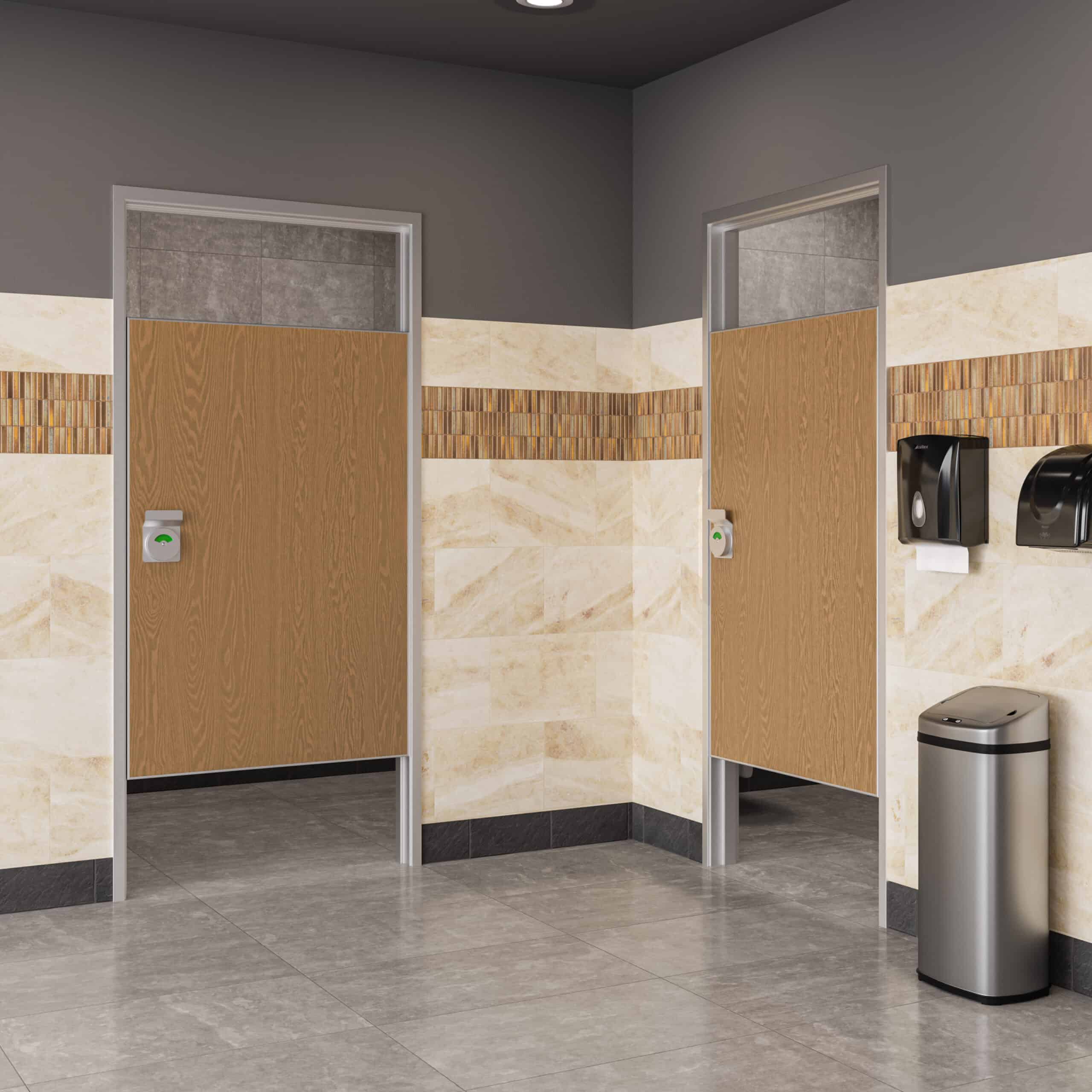 A public restroom with two stalls featuring partial wooden doors set in FlexFit™ Field-Adjustable Interior Aluminum Door Frames, beige tiled walls, a trash can, and paper towel dispensers on the right wall.