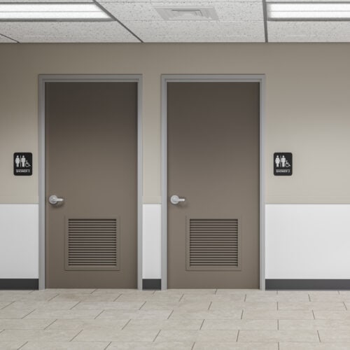 Two brown restroom doors in a hallway, each with a sign indicating accessible restrooms for all genders, are fitted with FlexFit™ Field-Adjustable Interior Aluminum Door Frames. The floor is tiled, and the walls are beige and white.