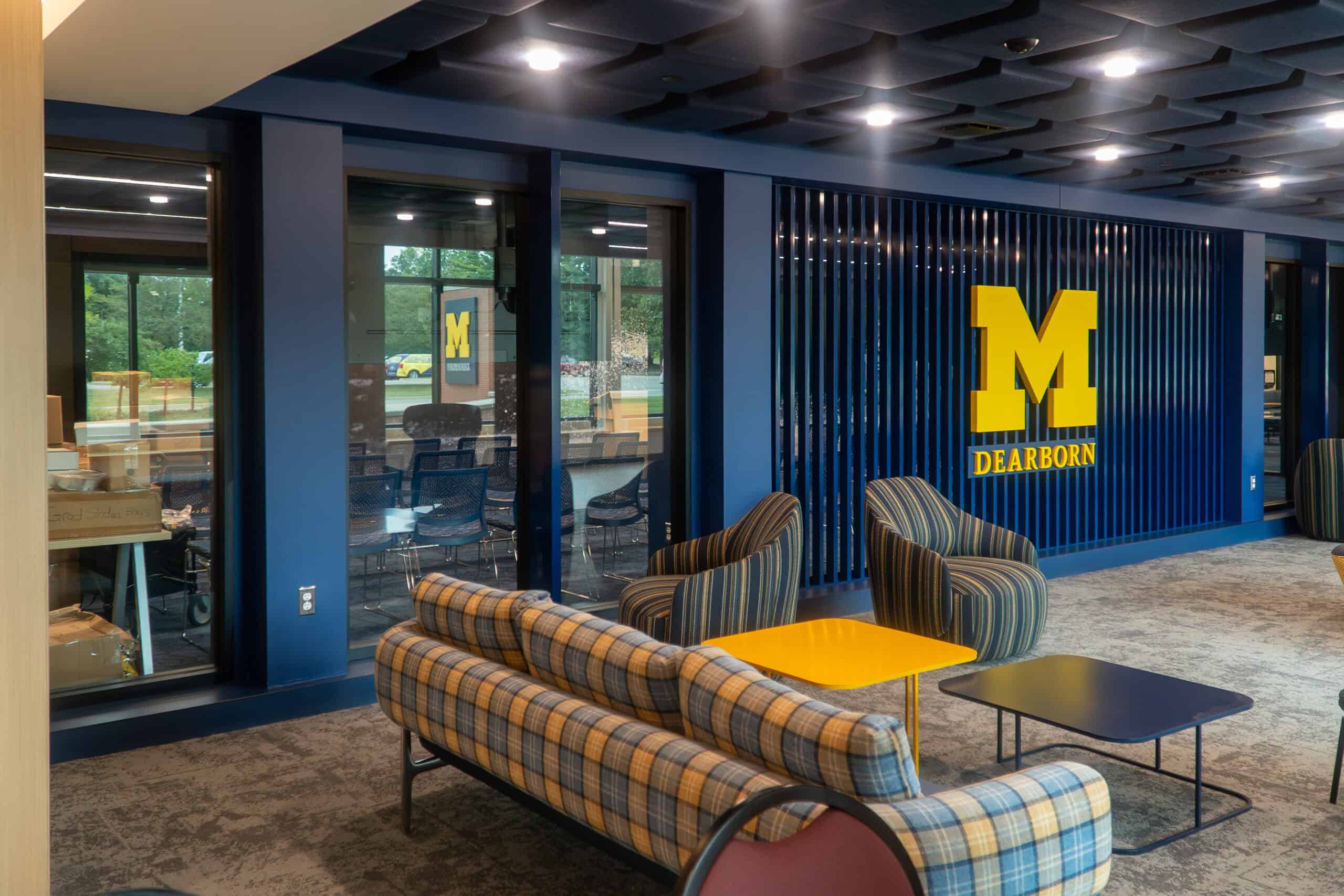 A modern lounge area with plaid sofas, striped chairs, and a yellow table creates an inviting hybrid work space, featuring a University of Michigan Dearborn logo on a blue wall.