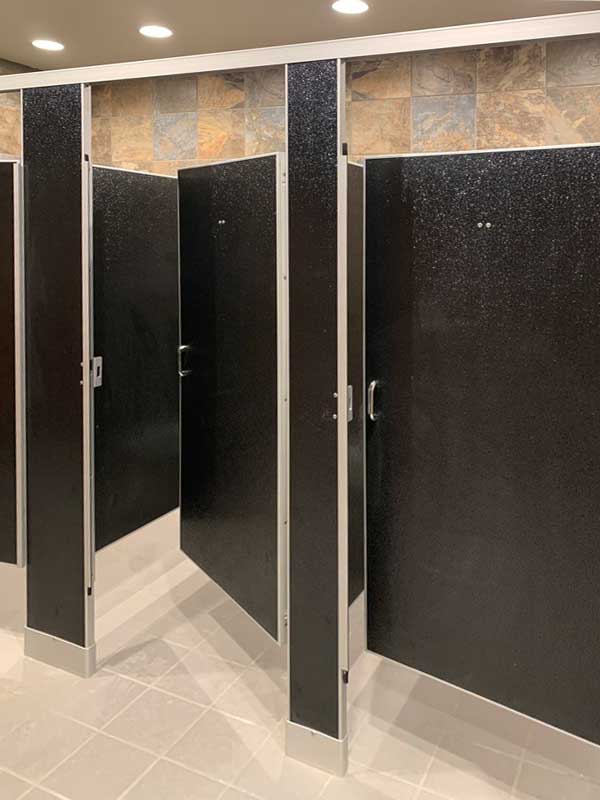 A row of black restroom stalls with doors slightly open, revealing empty stalls inside. The floor is tiled and the bright lighting reflects a clean environment, ideal for facilities focused on advanced healthcare solutions.