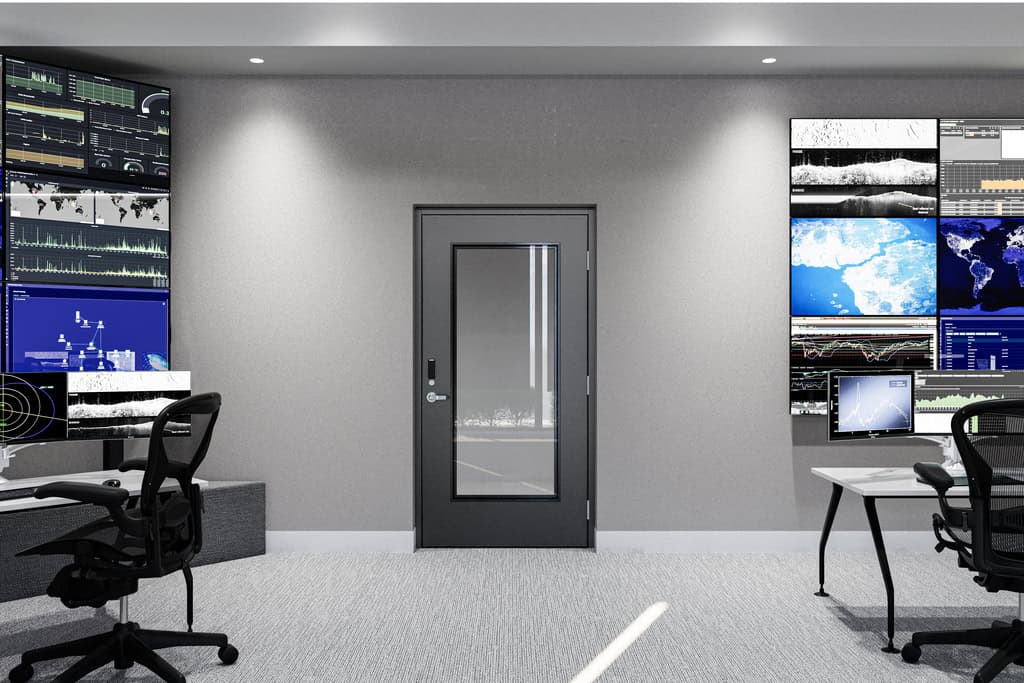 Modern control room with multiple wall-mounted monitors displaying data, maps, and graphs, two office chairs, desks, and a secure national account commercial door solution set in the center of a gray wall.