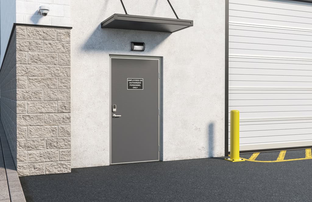 A gray metal door with a sign reading "Authorized Personnel Only" is set in a concrete wall next to a large garage door and a yellow bollard, showcasing secure entry typical of national account commercial door solutions.