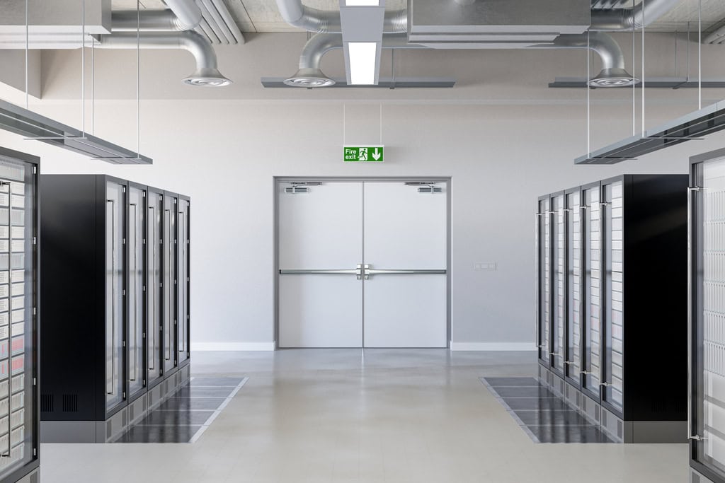 A modern data center aisle with server racks on both sides, overhead ventilation, and a double emergency exit door featuring national account commercial door solutions, marked by a green exit sign above.