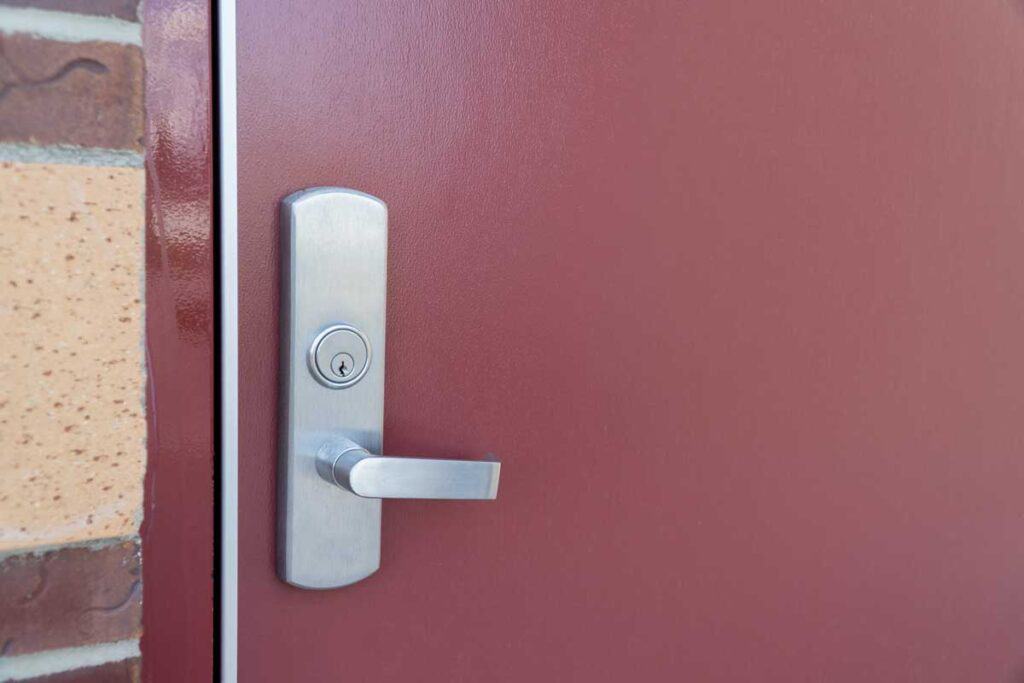 Close-up of a silver door handle and keyhole on a red door, with part of a brick wall visible—ideal for showcasing national account commercial door solutions.