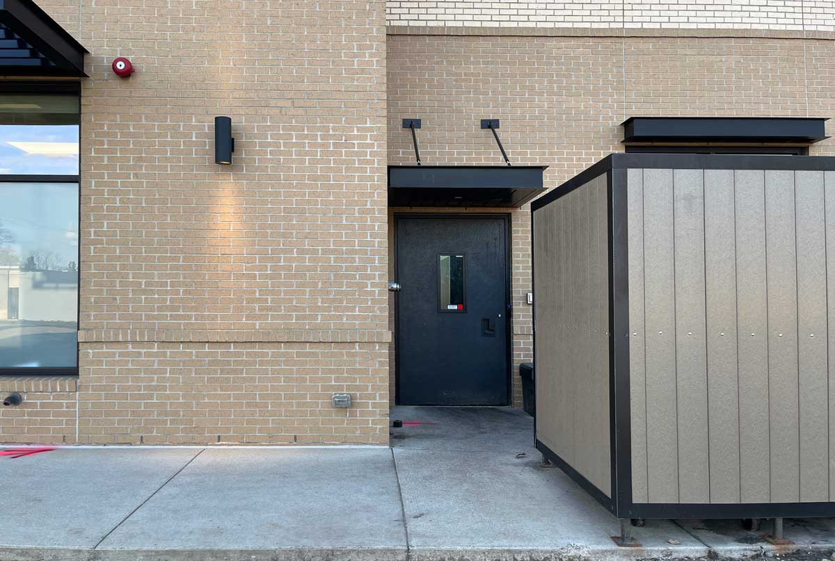 A closed black door on a beige brick building, partially obscured by a tall metal enclosure on the right, with a sidewalk in front—suggestive of secure access to modern healthcare solutions within.